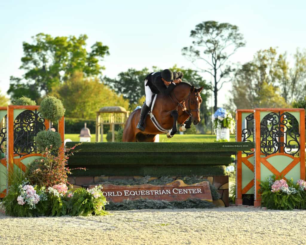 A rider and horse jump over an obstacle at an outdoor equestrian event, with "World Equestrian Center" displayed on a sign below the jump.