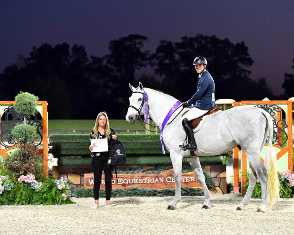 A rider on a white horse with a purple ribbon poses next to a woman holding an award certificate at an equestrian event.