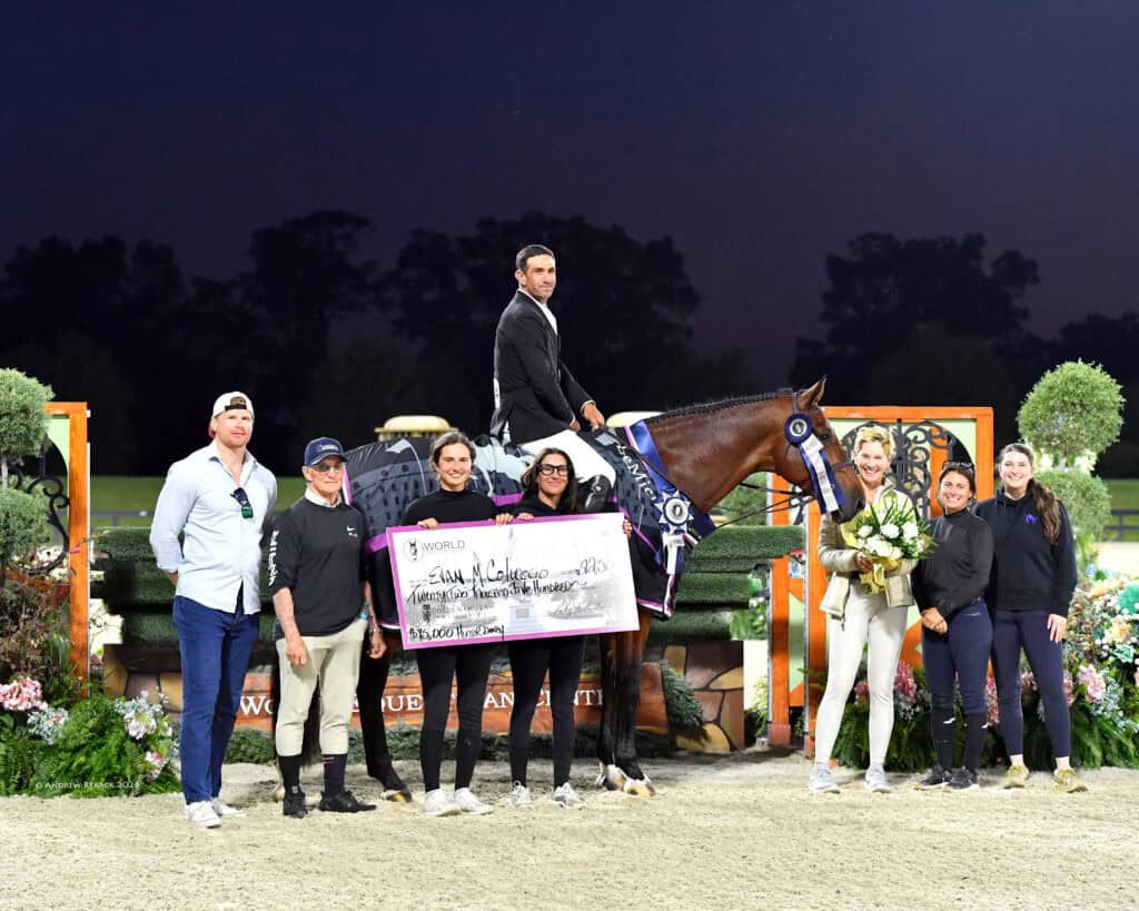 A group of people pose with a large ceremonial check; a man on horseback holds the check, while others stand nearby, some holding flowers, outdoors at dusk.