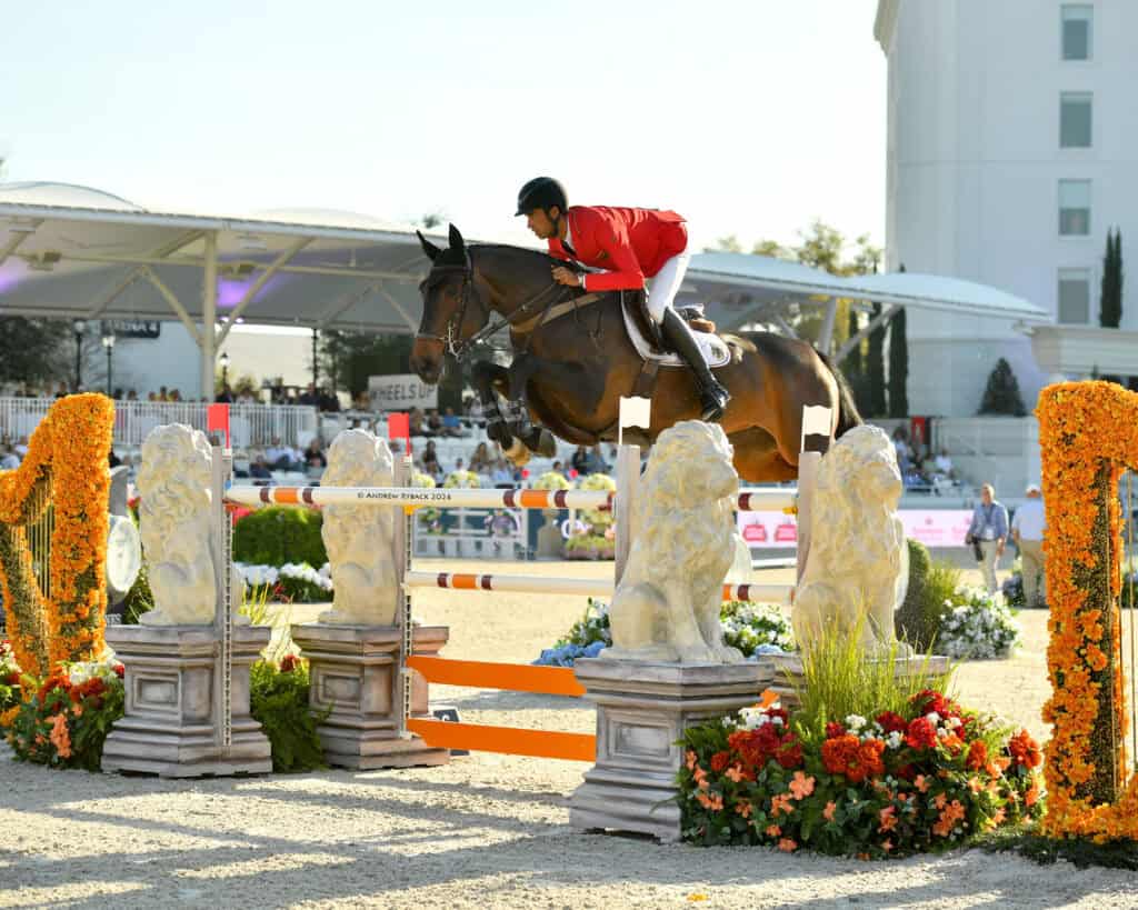 A rider in a red jacket and black helmet jumps a horse over an obstacle decorated with lion statues and flowers during an equestrian event.