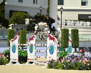 An equestrian rider in formal attire jumps a white horse over a colorful obstacle during a show jumping competition in an outdoor arena.