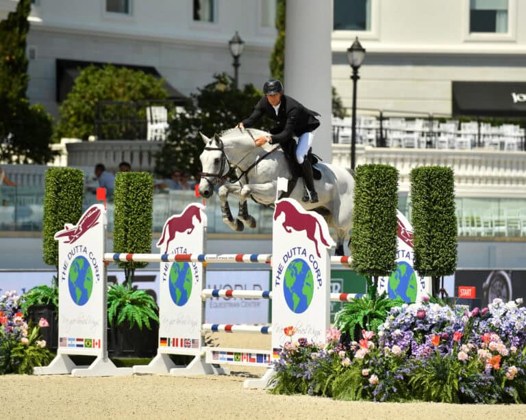 An equestrian rider in formal attire jumps a white horse over a colorful obstacle during a show jumping competition in an outdoor arena.