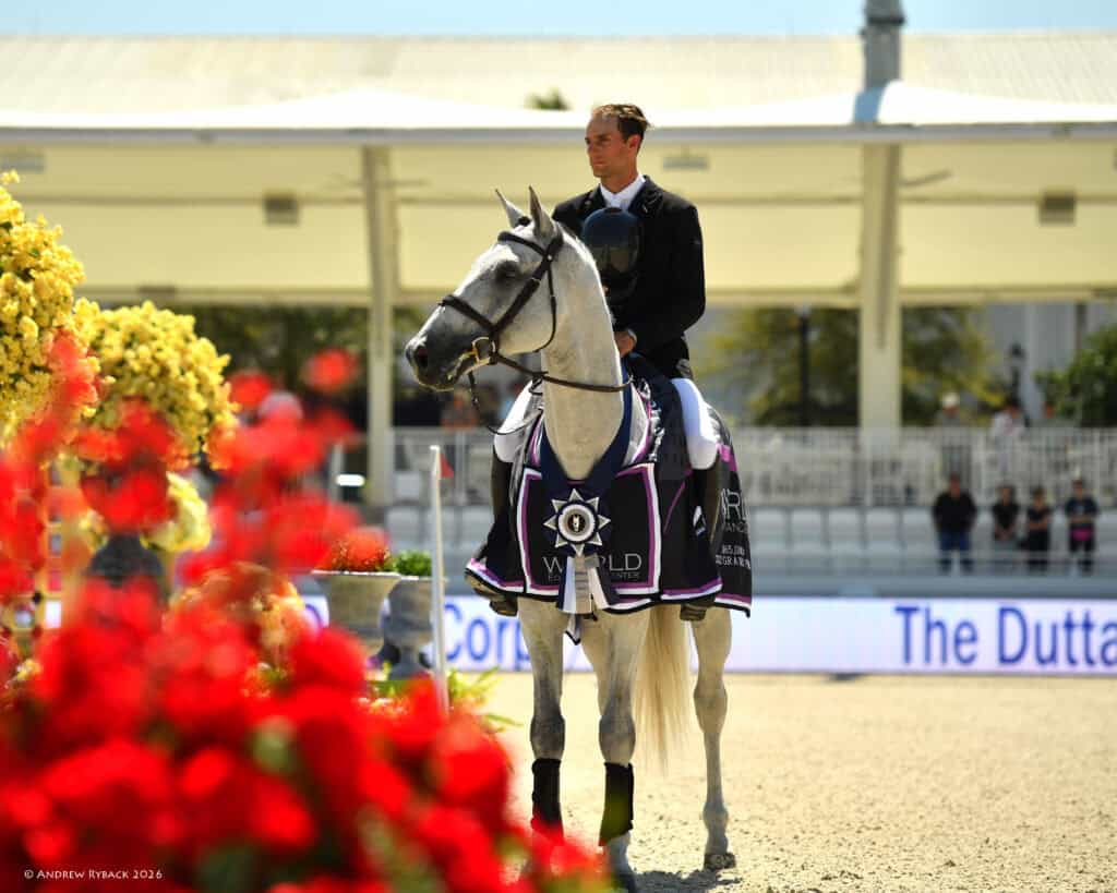 A rider in formal attire sits on a gray horse draped in a winner's blanket, surrounded by flowers, in an outdoor equestrian arena.