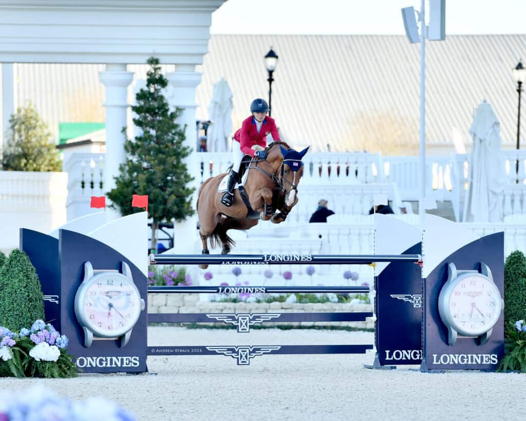A rider in a red jacket and helmet guides a horse over a high Longines-branded jump at an equestrian event, with a white pavilion in the background.