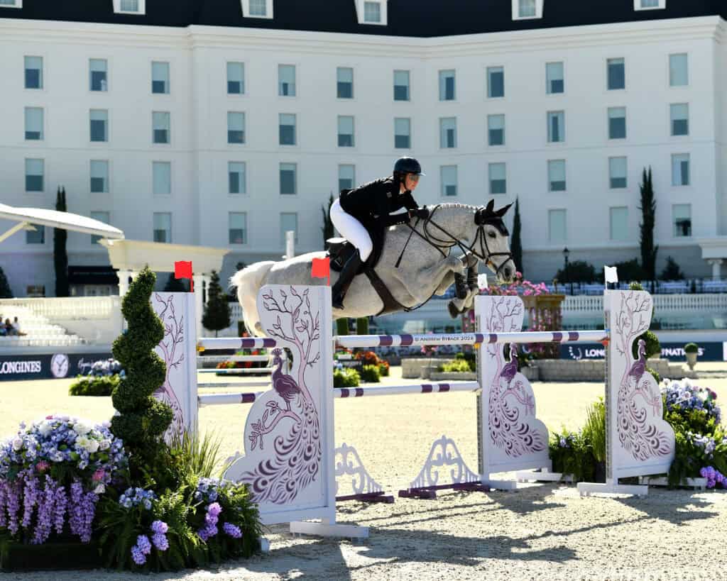 A rider on a gray horse jumps over an obstacle during an equestrian event, with a white building in the background and flower decorations around the course.