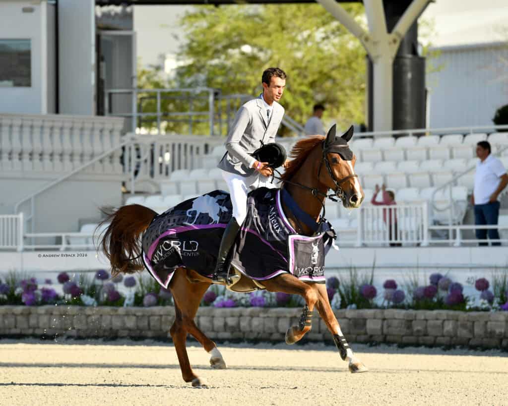A rider in a light grey jacket sits on a horse draped in "World" branded coverings, trotting in an outdoor arena with empty white stands in the background.
