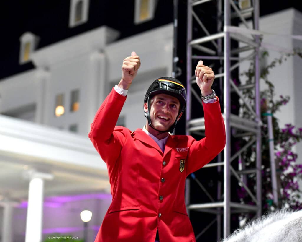 A male equestrian in a red jacket and helmet raises both thumbs up, smiling, with a building and event lighting in the background.