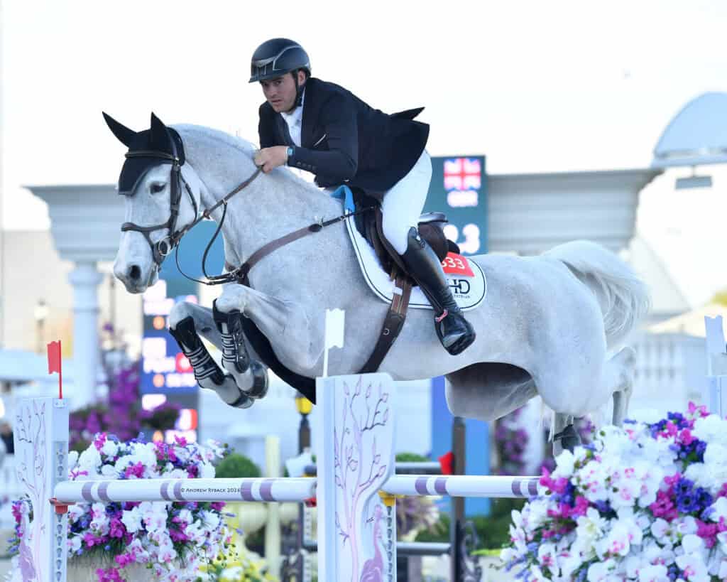A rider in formal attire jumps a gray horse over a colorful obstacle during a show jumping competition.