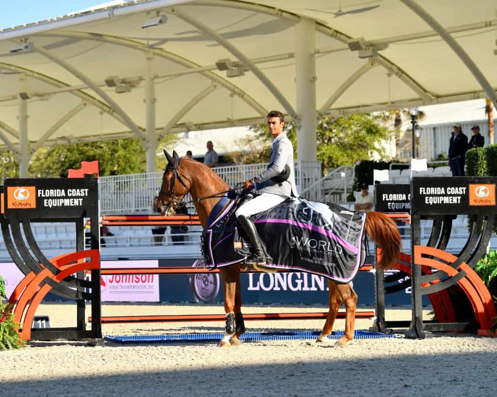 A rider in competition attire sits on a horse draped in a branded blanket, positioned in an arena near two showjumping obstacles and spectator seating.