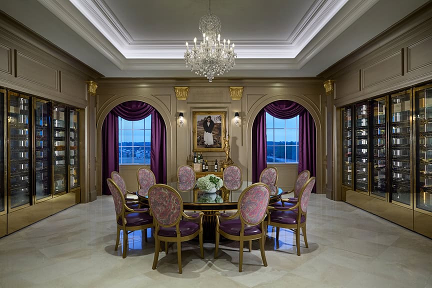 Elegant dining room with a glass table, eight ornate chairs, chandelier, bookshelves, large windows with purple curtains, and a portrait above a fireplace mantel.