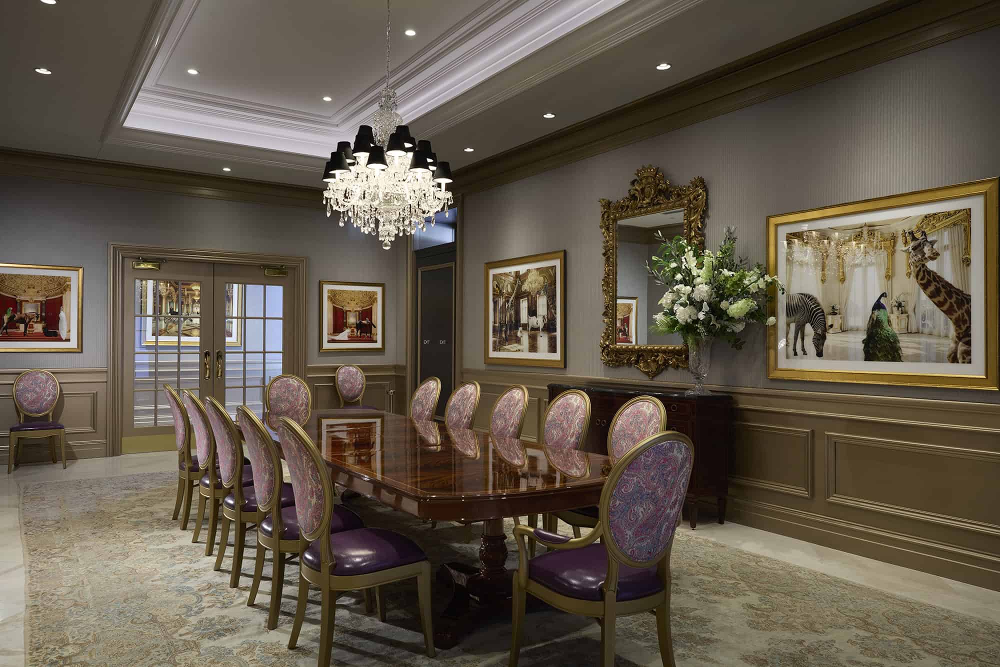 Elegant conference room with a polished wooden table, ornate chandelier, purple-upholstered chairs, large mirror, framed artwork, and floral arrangement on a patterned rug.