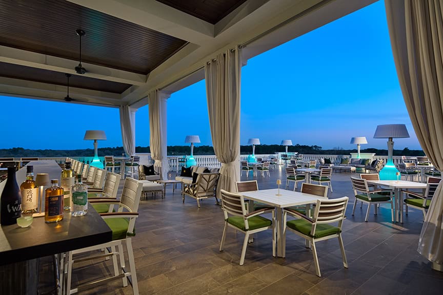 Outdoor terrace bar with white tables and chairs, bottles on the counter, glowing lamps, and a view of the evening sky.