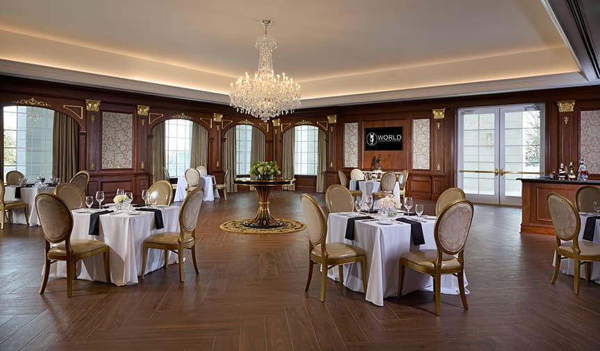 Elegant dining room with round tables, gold chairs, chandelier, and wood-paneled walls; set for a formal event with place settings and floral centerpieces.
