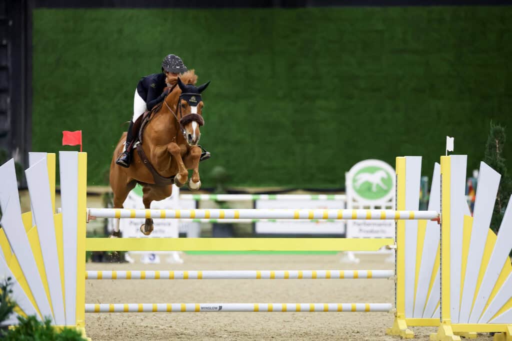 A rider and horse jump over a yellow and white obstacle during an indoor equestrian show jumping competition.