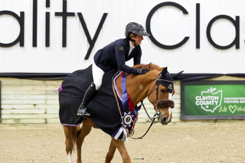Equestrian rider in formal attire sits on a decorated horse wearing a winner's sash and ribbons at an indoor competition venue.