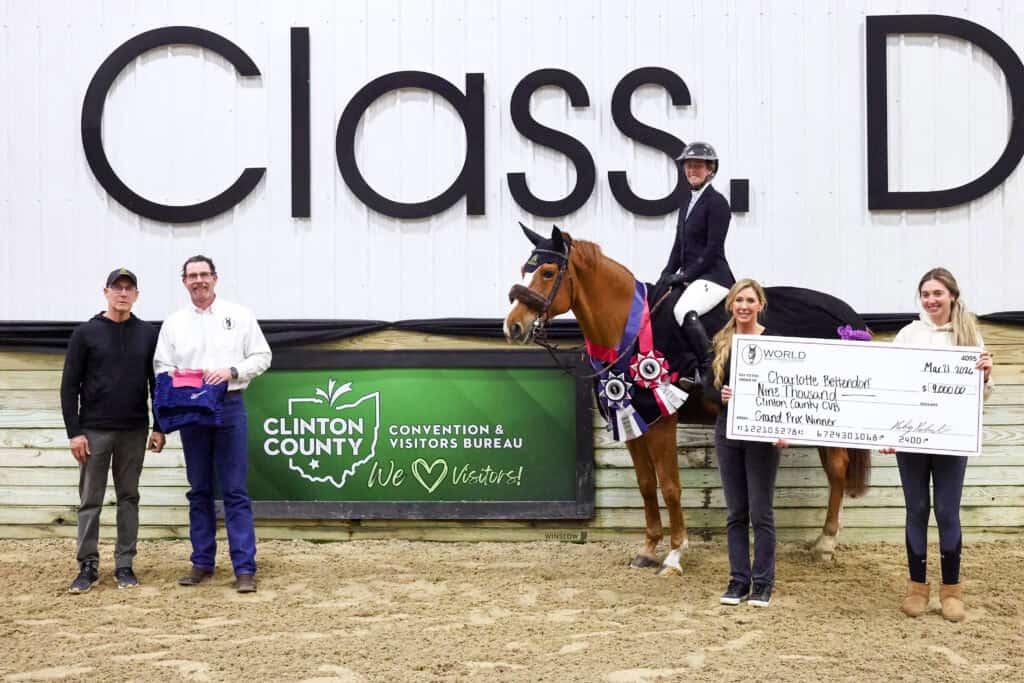 Five people and a horse pose indoors; one person on horseback wears riding attire and ribbons, while two others hold a large ceremonial check and prize items in front of a Clinton County sign.