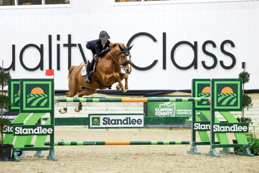 Equestrian rider and horse jump over a green and orange obstacle during an indoor show jumping competition.