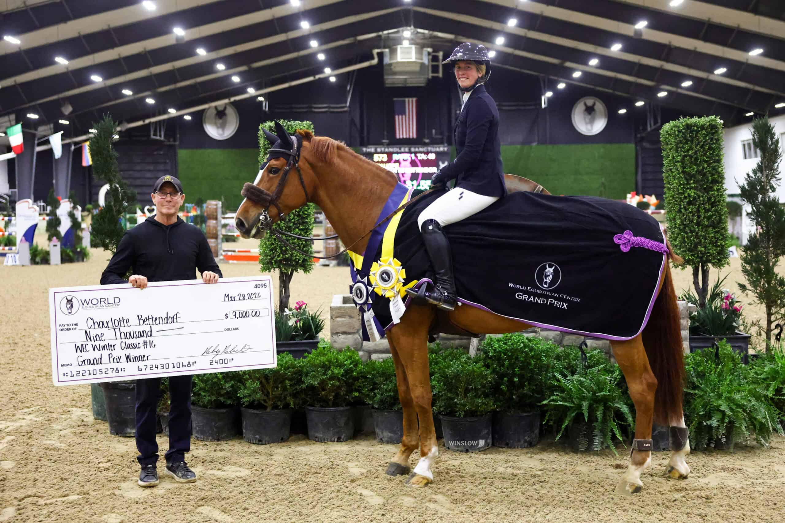 A rider on a horse with a winner's sash poses in an indoor arena next to a person holding a large ceremonial check.