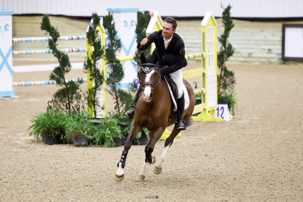 A rider in formal attire gives a thumbs-up while riding a brown and white horse on an indoor show jumping course.