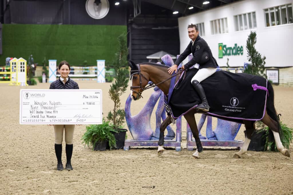 A woman holds a large presentation check while a man in riding attire sits on a horse draped with a winner’s blanket at an indoor equestrian event.