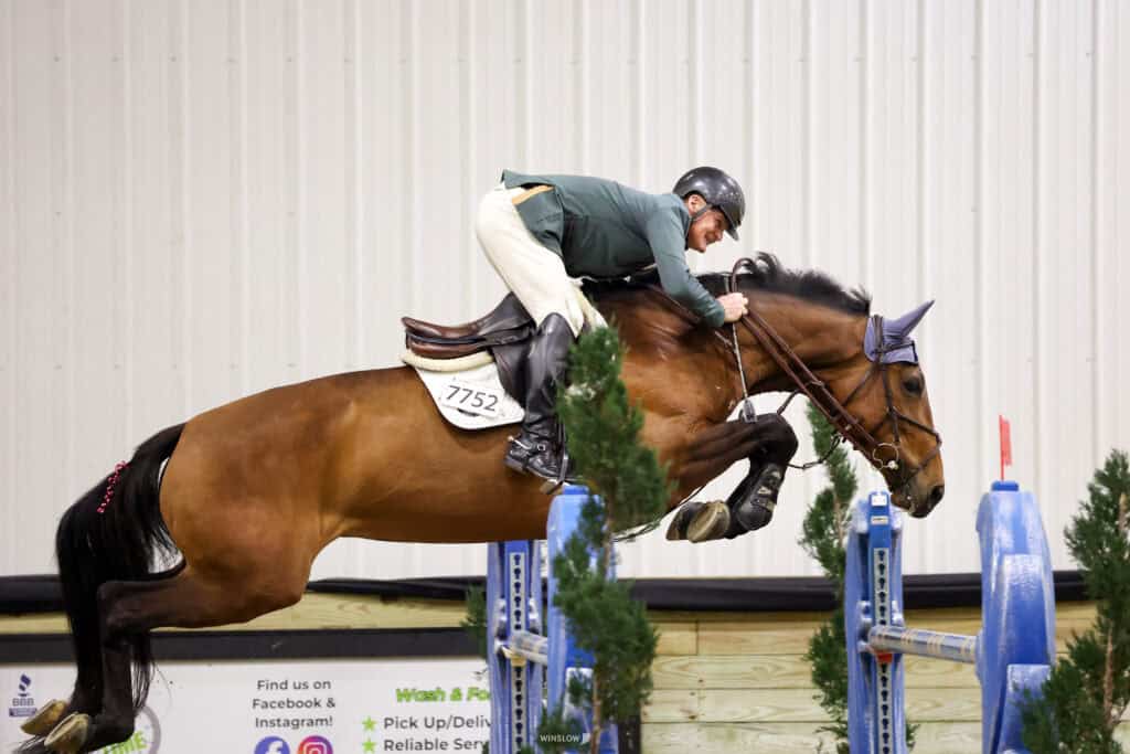 A rider in show jumping attire guides a horse over a blue and white obstacle during an indoor equestrian event.