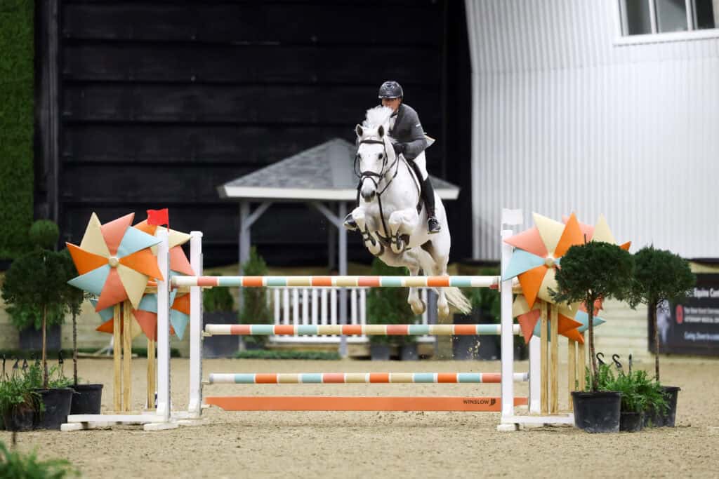 A rider in equestrian gear jumps a white horse over a colorful obstacle during an indoor show jumping competition.