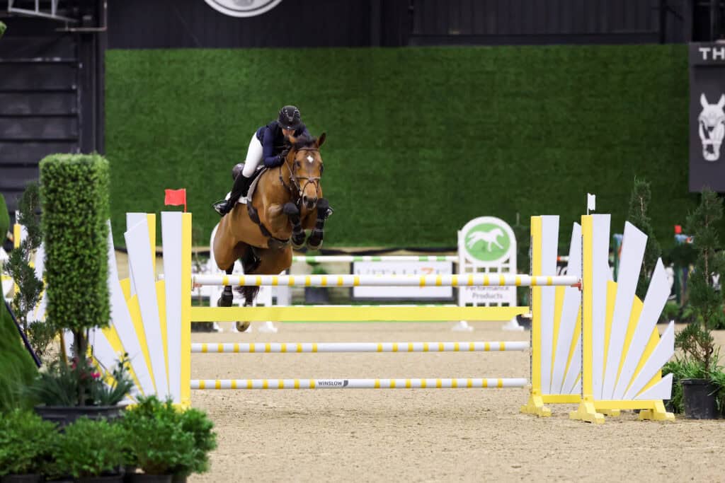 A rider in equestrian gear and helmet jumps a brown horse over a white and yellow hurdle during an indoor show jumping competition.