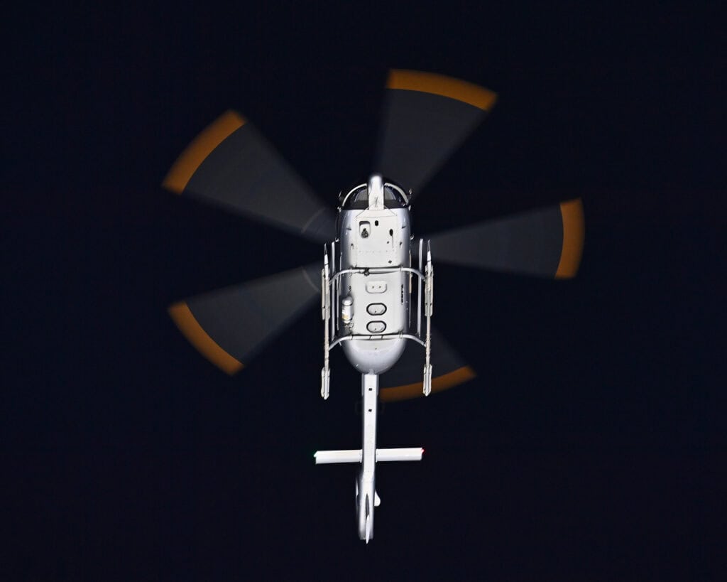 A helicopter flying at night, viewed from below, with its rotor blades in motion against a dark background.