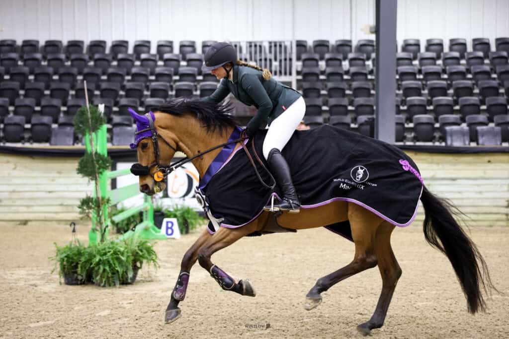 A rider in a helmet and equestrian attire guides a horse in full tack and a black blanket across an indoor arena with empty bleachers in the background.