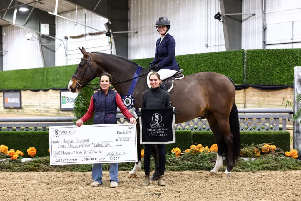 Three people at an indoor equestrian event: one on horseback, two standing and holding a large check. The setting includes greenery and autumn decorations.