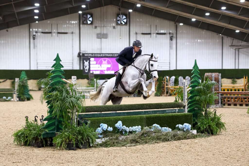 A person riding a white horse jumps over a green obstacle during an indoor equestrian event.