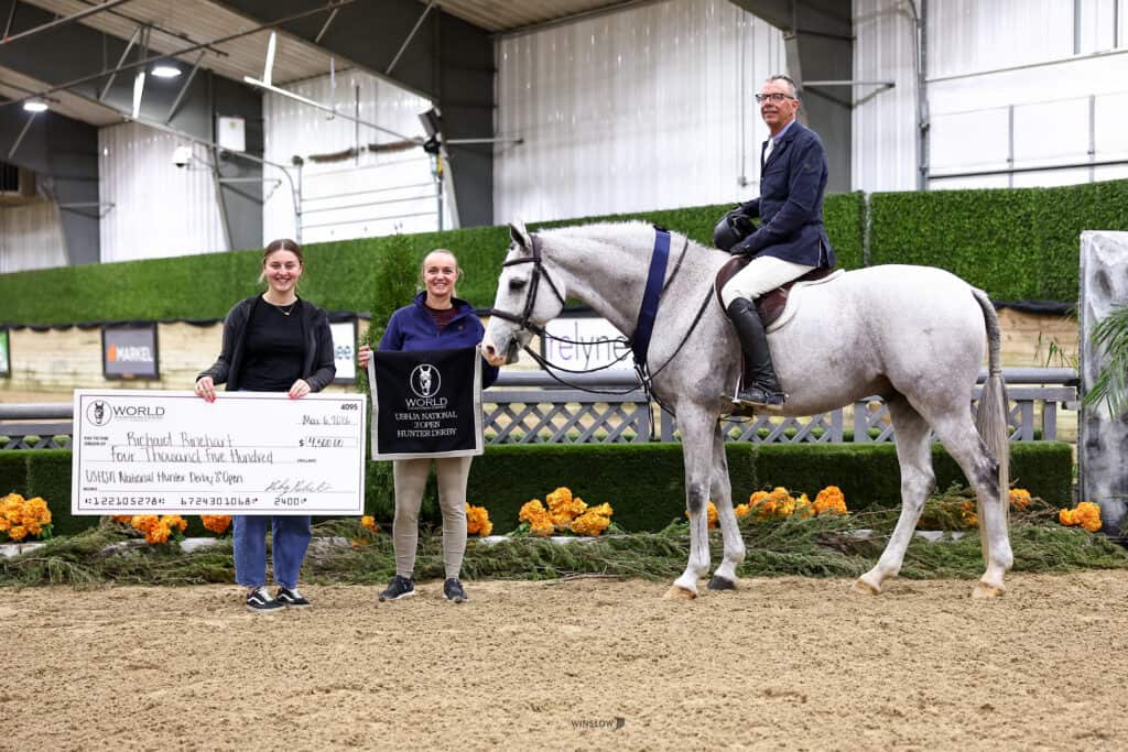 Three people pose in an indoor arena with a white horse; one holds a large check, another holds a plaque, and the third is mounted on the horse.