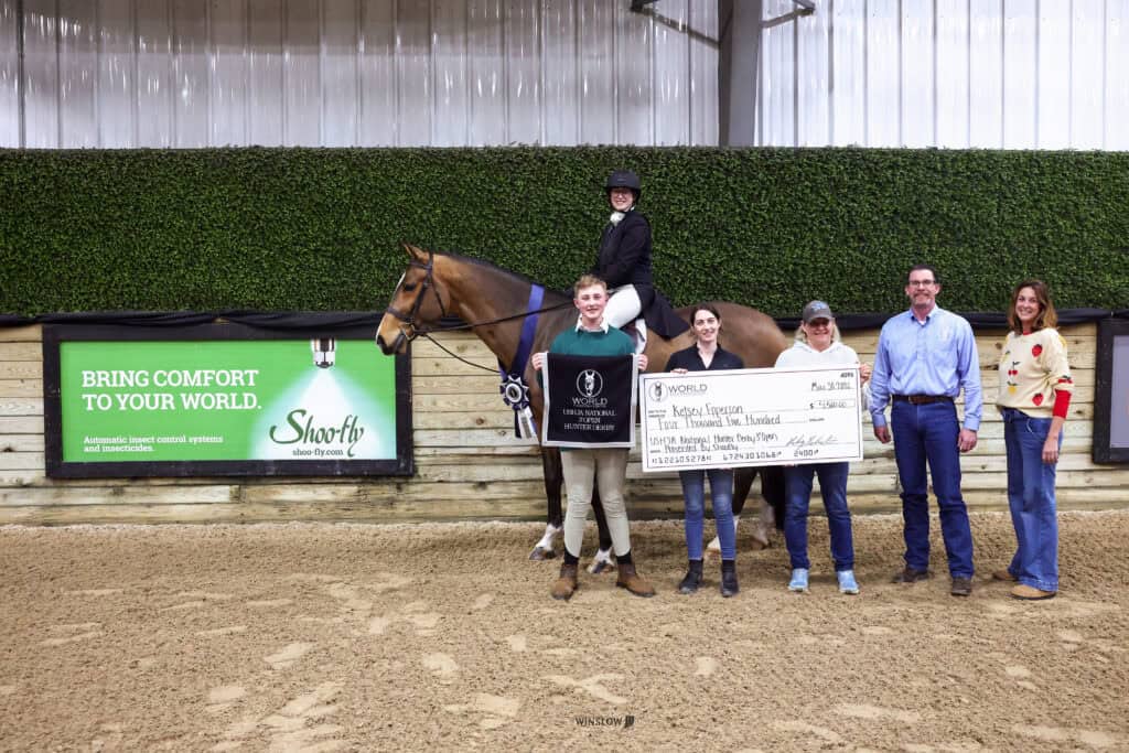 A group of six people, including one person on horseback, pose indoors with a large prize check and a ribbon in front of a green hedge and advertisement signs.