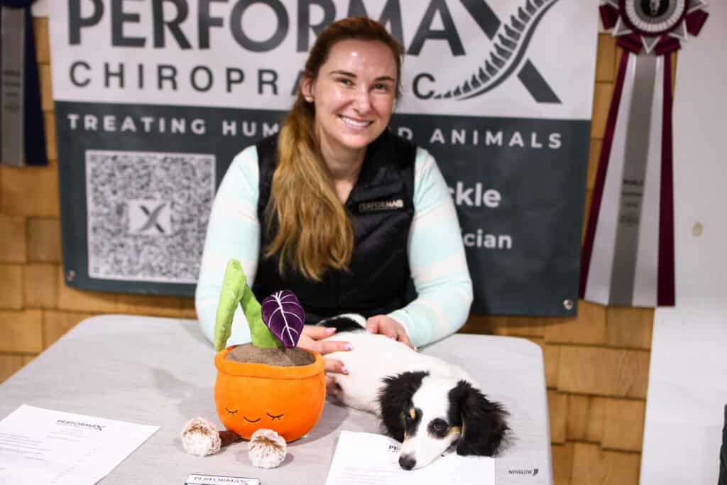A woman sits at a booth with a dog on the table, promotional materials, a plant plush, and a Performax Chiropractic banner in the background.