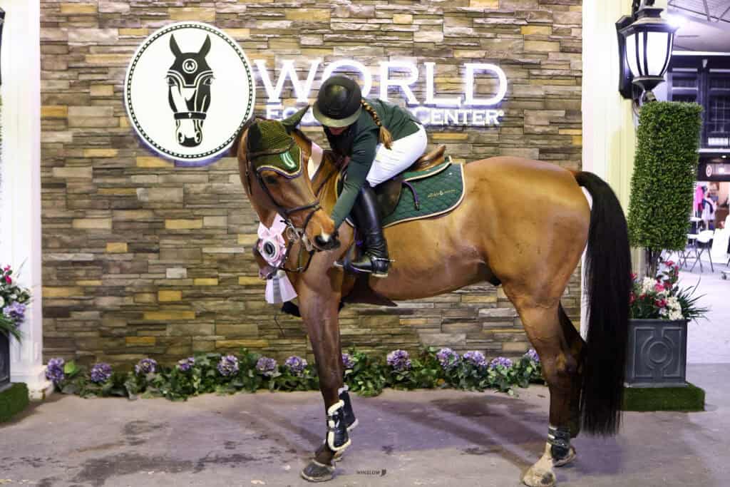 A rider in equestrian attire hugs a horse wearing a green saddle pad and a ribbon, standing in front of a “World Equestrian Center” sign with a stone wall backdrop.