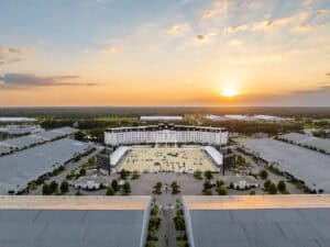 Aerial view of a large equestrian facility with a central arena surrounded by grandstands and buildings at sunset.