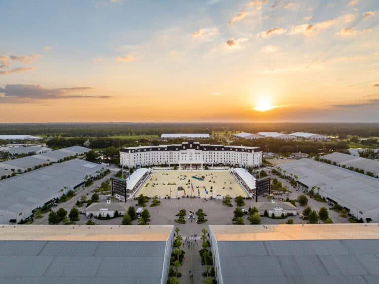 Aerial view of a large equestrian facility with a central arena surrounded by grandstands and buildings at sunset.