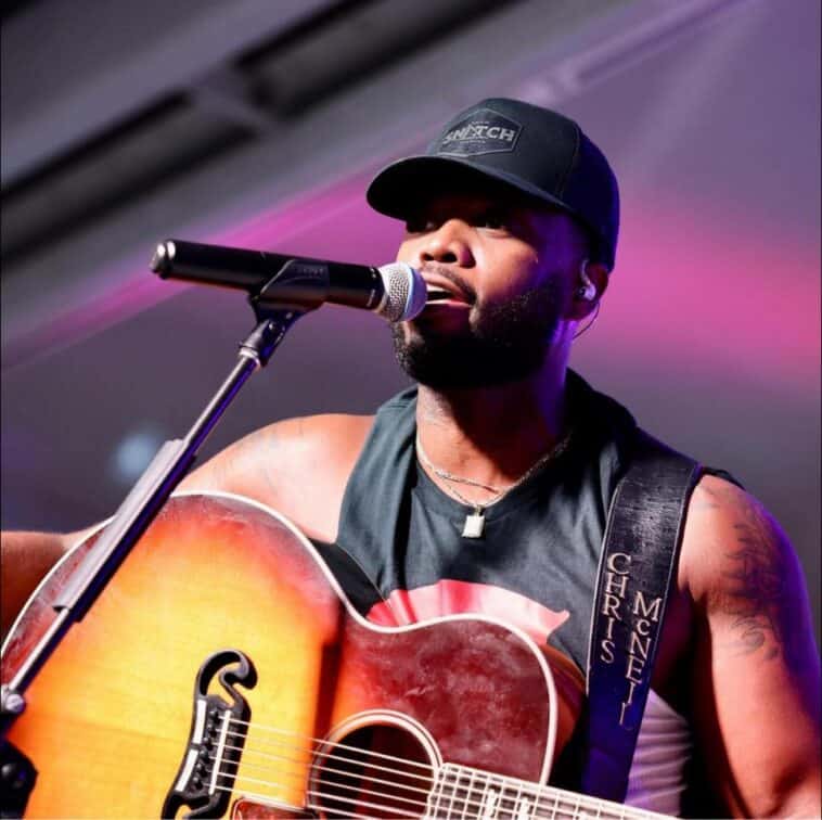 A man wearing a black cap and sleeveless shirt plays an acoustic guitar and sings into a microphone on stage.