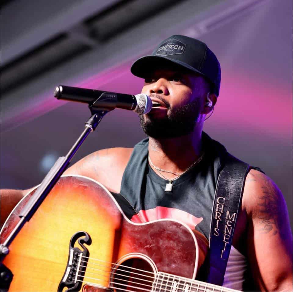 A man wearing a black cap and sleeveless shirt plays an acoustic guitar and sings into a microphone on stage.