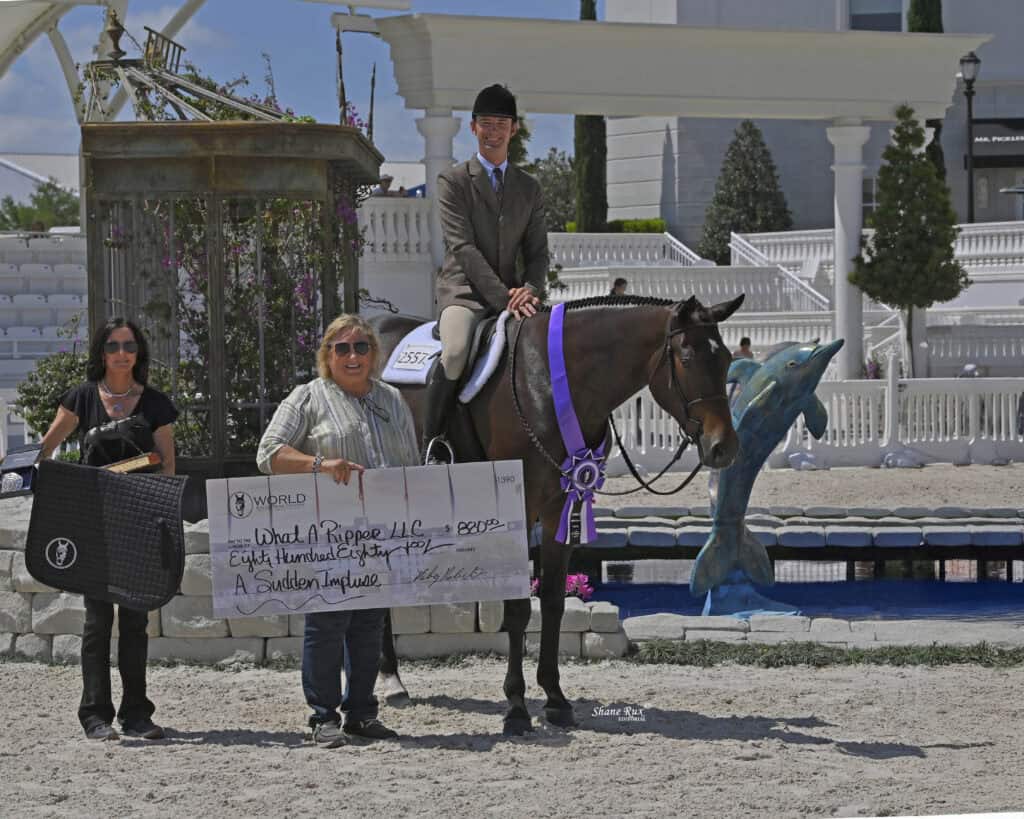 Three people and a horse pose outdoors; one person on horseback with a ribbon, two holding a large check, and equestrian equipment nearby. Fountain and white building in background.