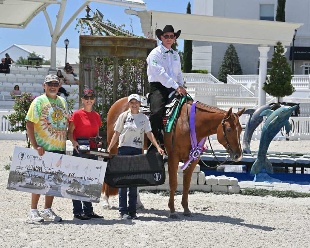 Four people and a horse pose outdoors; one person holds a large check, another holds an award case, and the rider on the horse wears a hat and medals. Decorative dolphins are in the background.