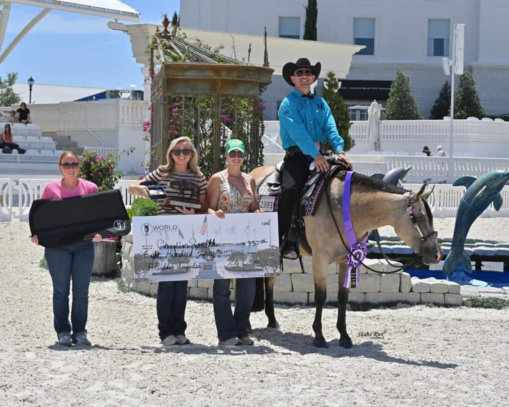 Four people and a horse pose outdoors with a large check, ribbon, and a prize case at an equestrian event.
