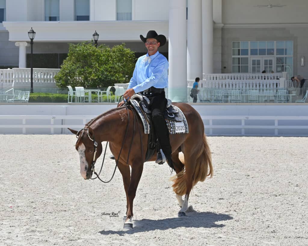 A man in a cowboy hat and blue shirt rides a brown horse with a braided mane in an outdoor arena near a white building.