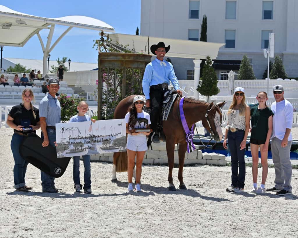 A group of people pose outdoors with a horse and rider, large check, guitar case, and ribbons, likely at an equestrian event or competition.