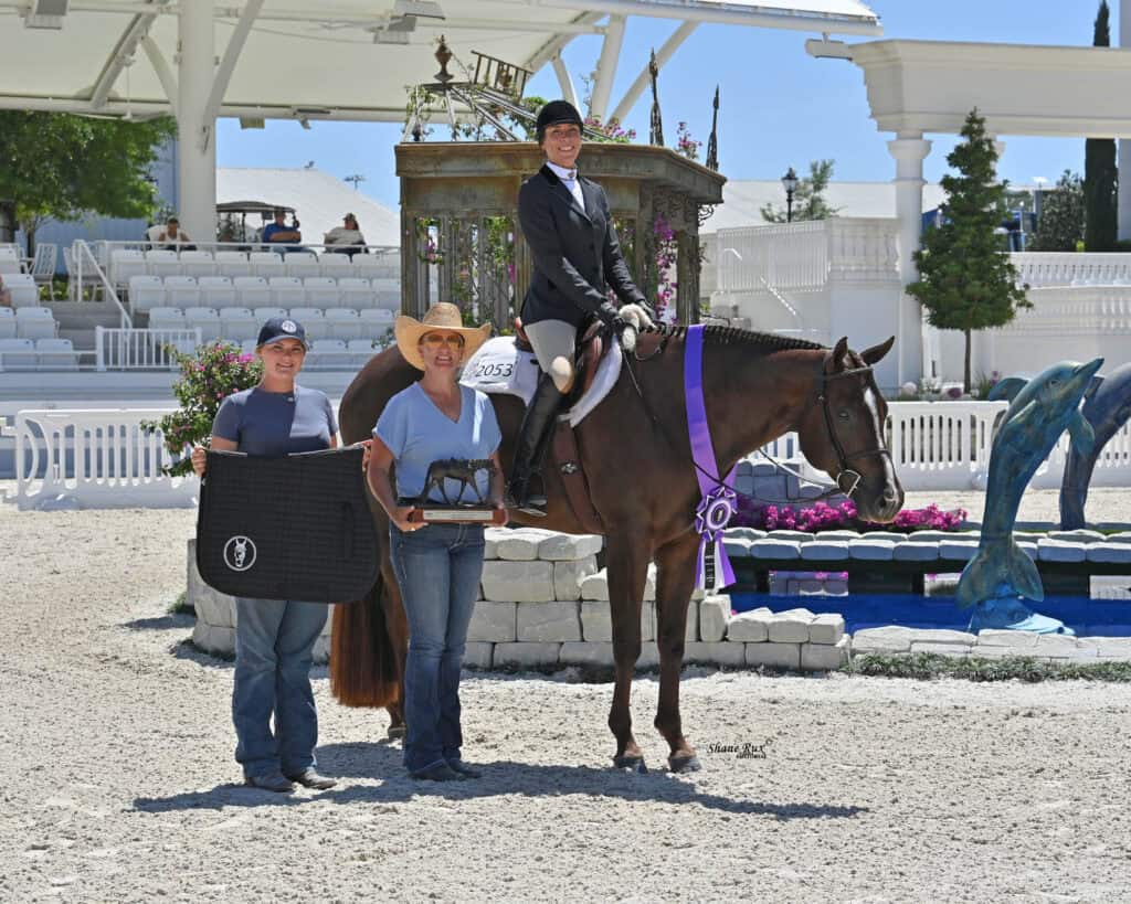 Three people at an equestrian event: one on horseback with a ribbon, and two standing, holding a saddle pad and trophy. An arena with seating and decorations is in the background.