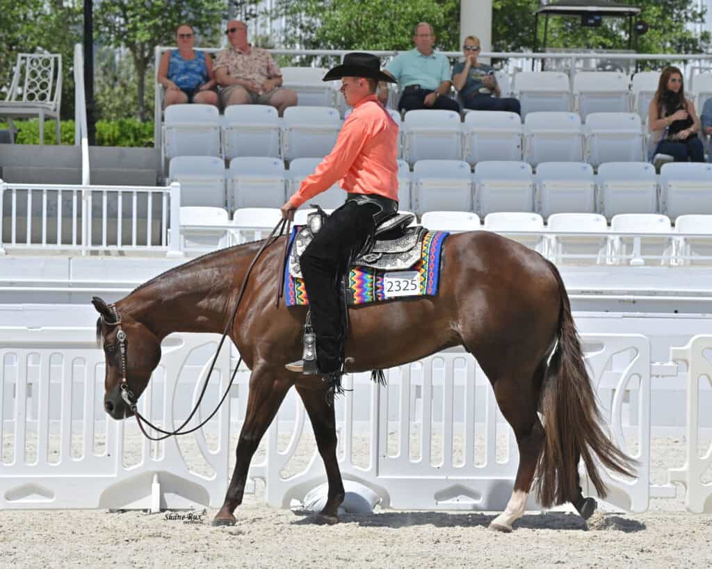 A man in a black cowboy hat and orange shirt rides a brown horse with a colorful saddle pad in an outdoor arena; spectators are seated in the background.