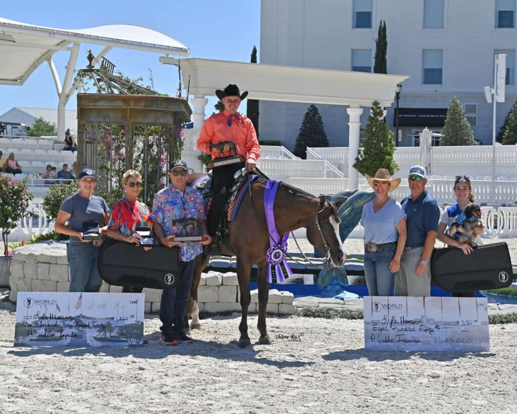 A group of people, including a person on horseback with a purple ribbon, pose outdoors with large checks and cases at an equestrian event.
