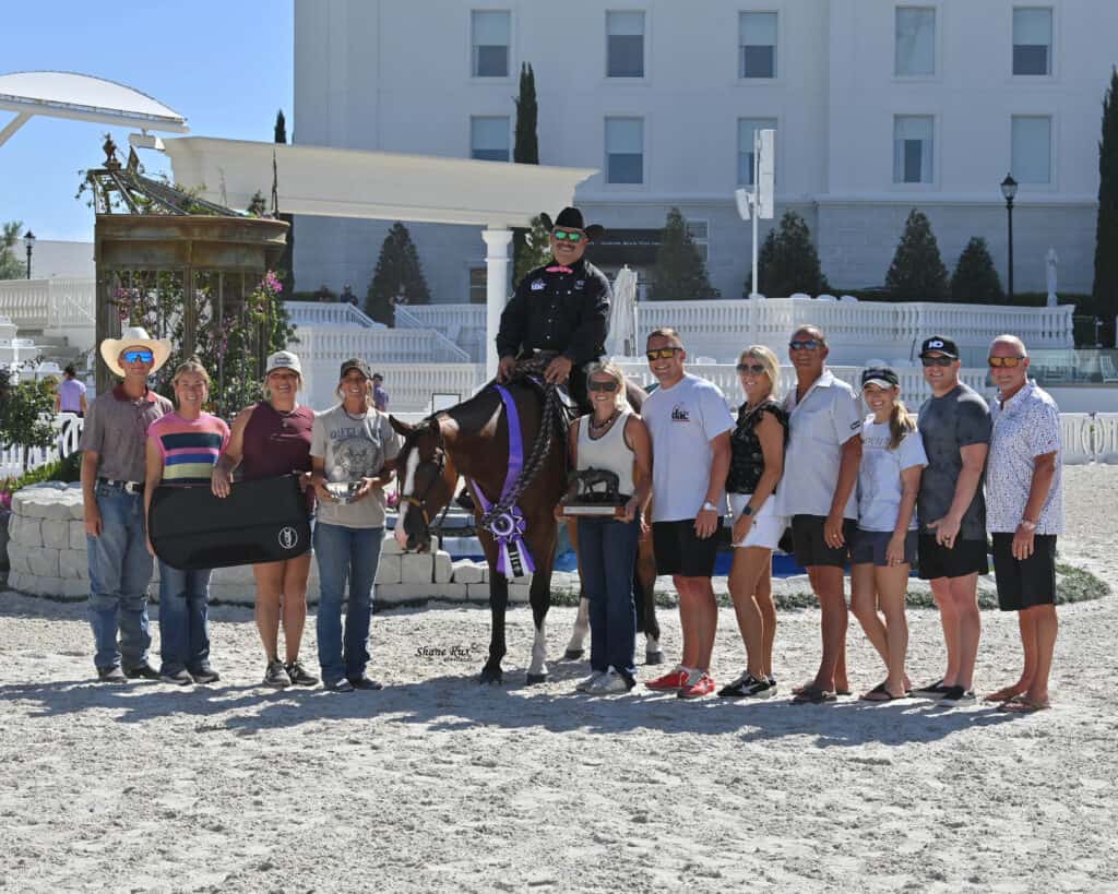 A group of people pose outdoors with a person on horseback wearing a ribbon and holding trophies. The setting includes a white building and decorative elements in the background.