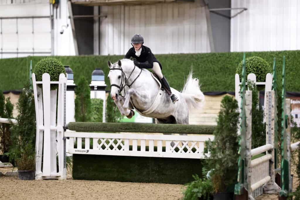 A rider on a gray horse jumps over a green and white obstacle during an indoor equestrian event.