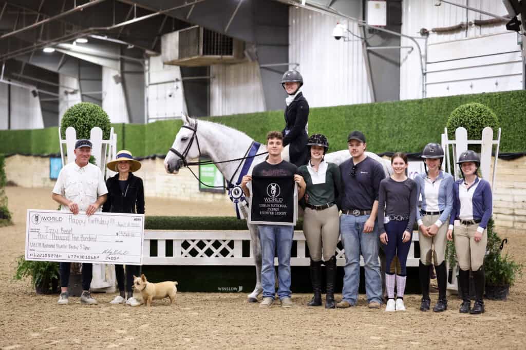 A group of people poses indoors with a white horse and rider; one person holds an oversized check. A small dog stands in front of the group.